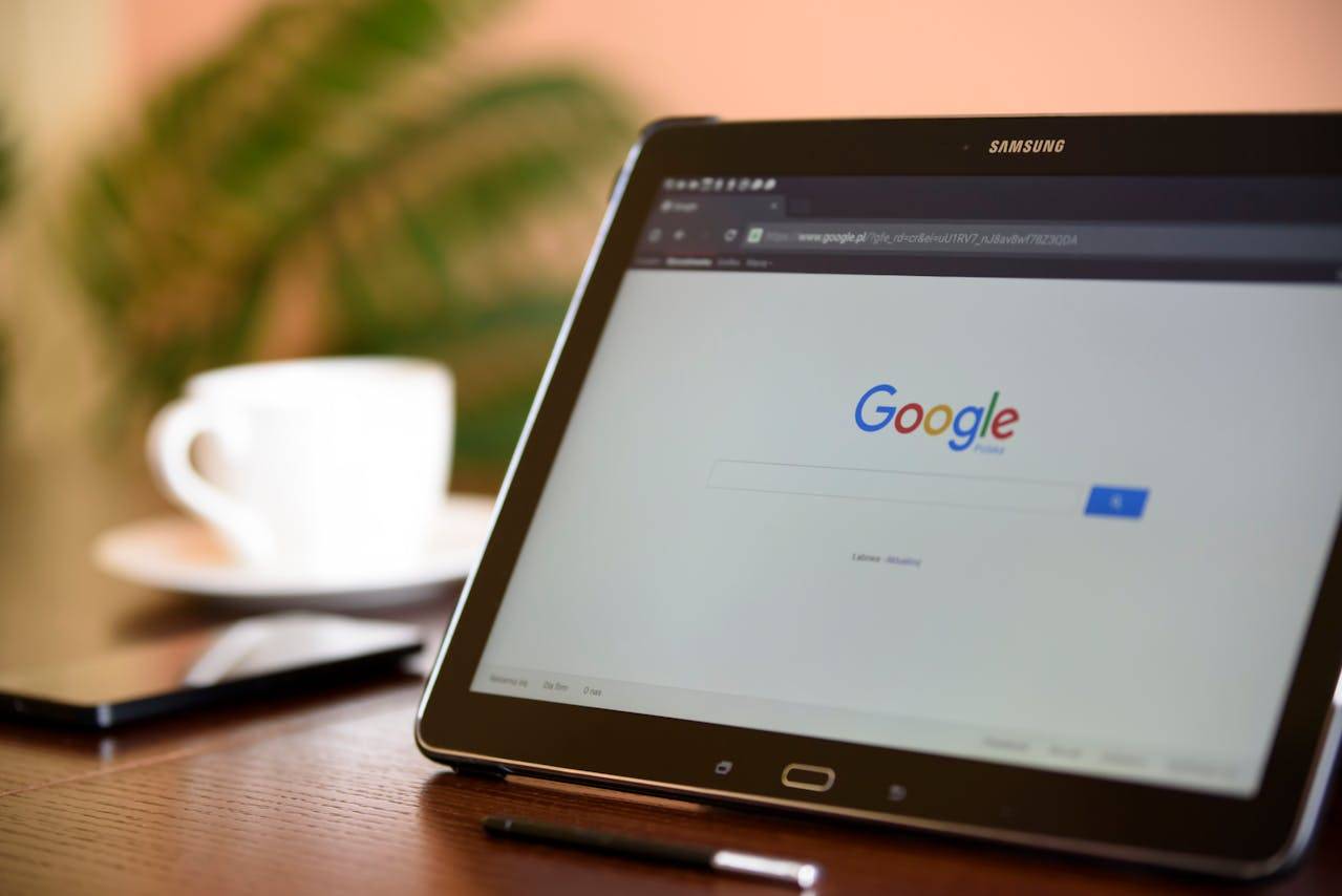 Electronic tablet with Samsung branding on the screen, next to a coffee cup on a saucer. Text on the screen reads: "www.google.pl/pferd-cr&ei=uUTRV7 Bav8wf7823QDA Google".